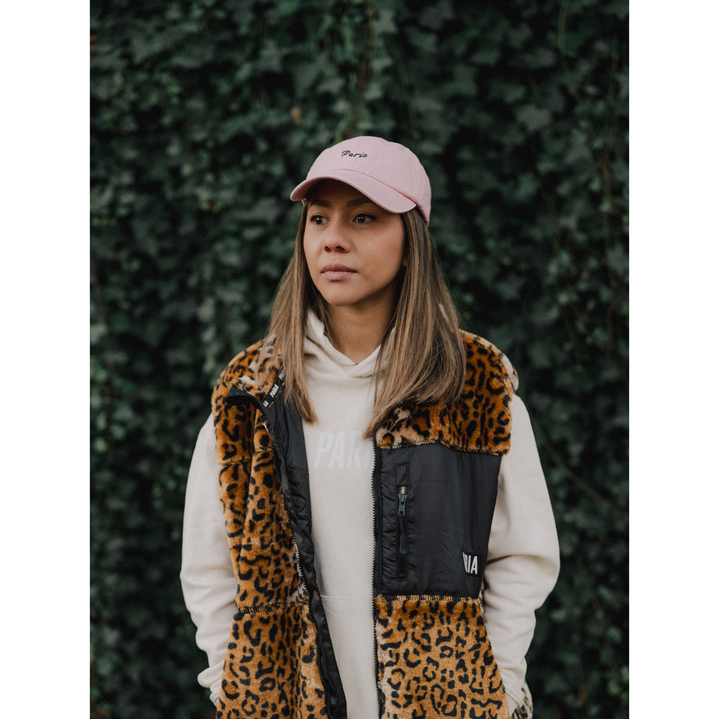 Person wearing a leopard print vest and pink cap against a green hedge background