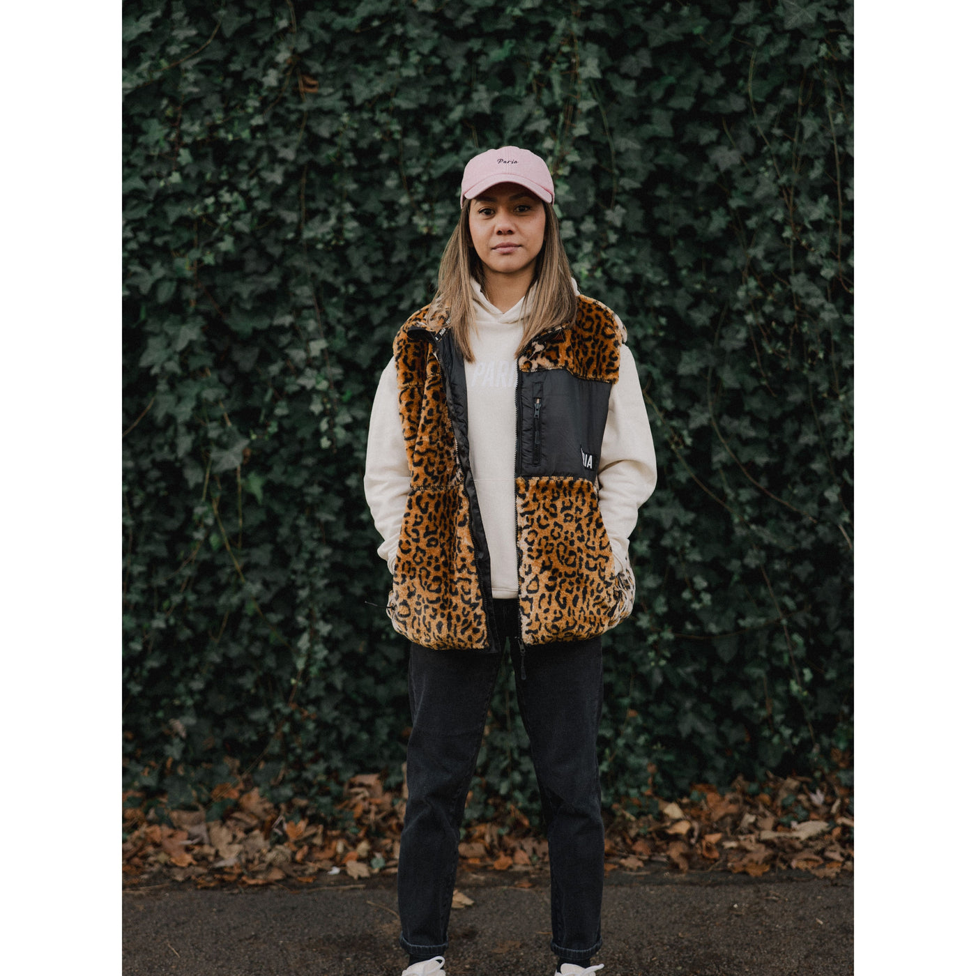 Person wearing a leopard print vest and pink cap against a green ivy wall.