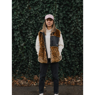 Person wearing a leopard print vest and pink cap against a green ivy wall.