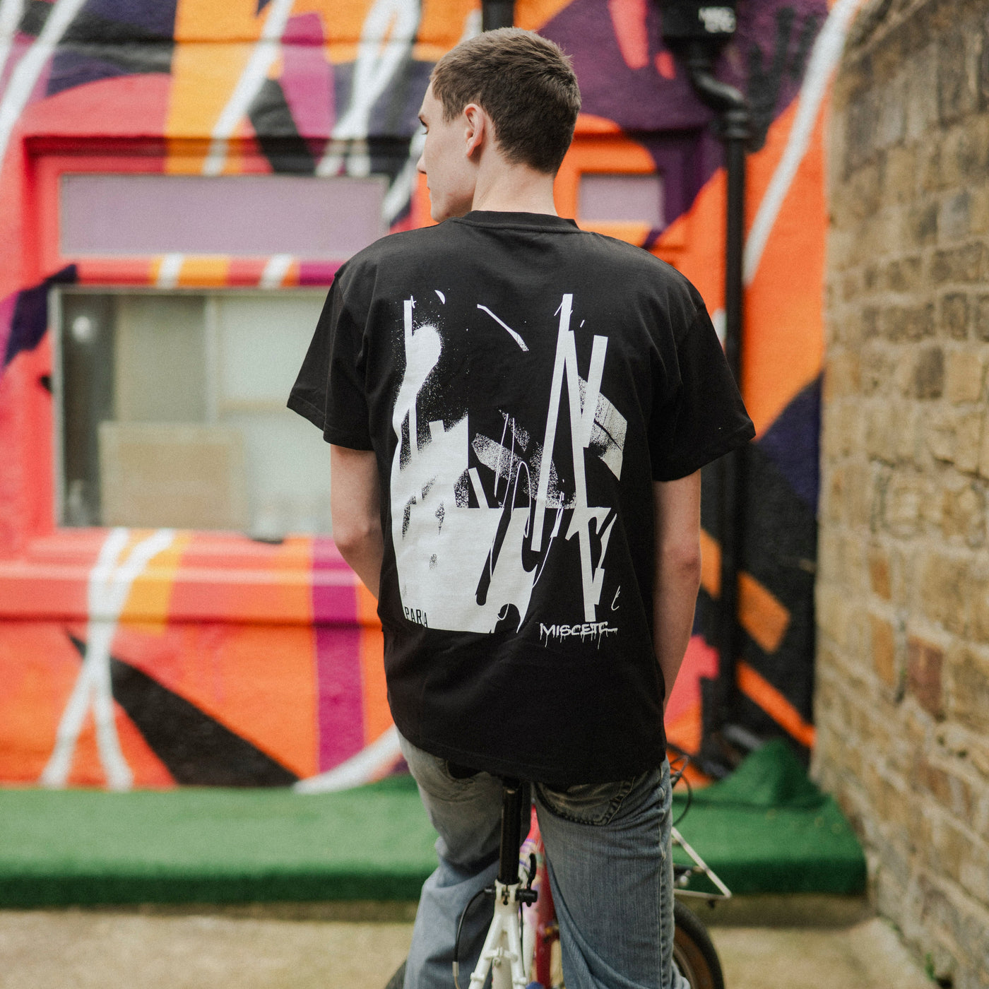 Person on a bicycle wearing a black t-shirt with white graphics in front of a colorful mural.