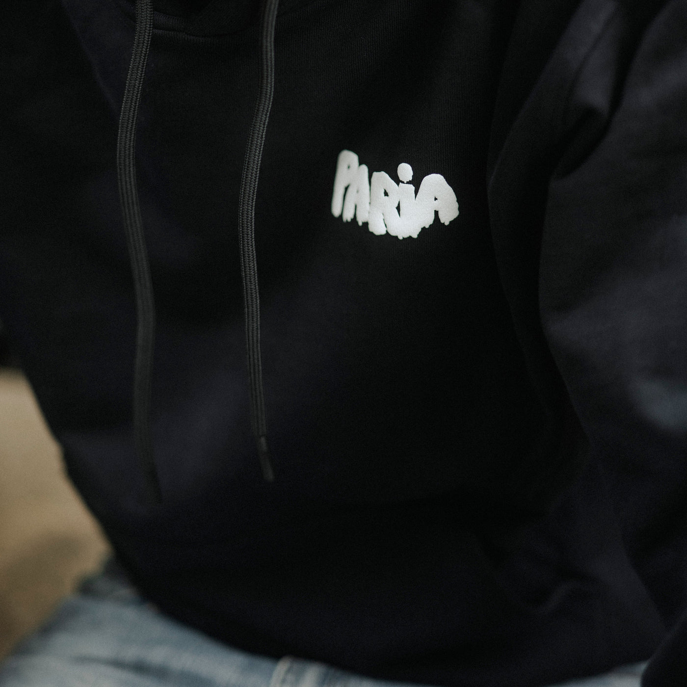 Person wearing a black hoodie with 'Paria' embroidered on it, blurred background