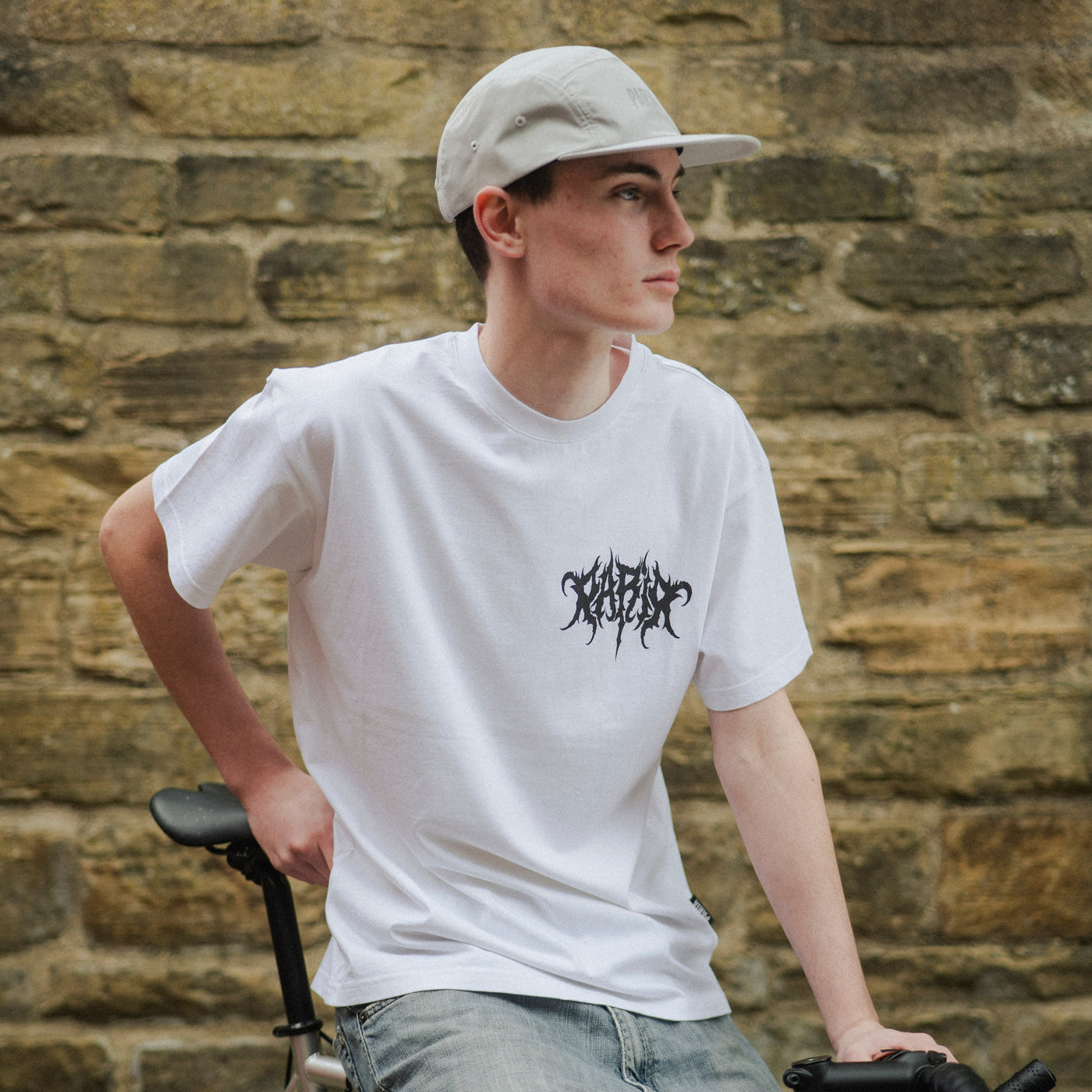 Person wearing a white t-shirt with black text and a cap, standing next to a bicycle against a brick wall.