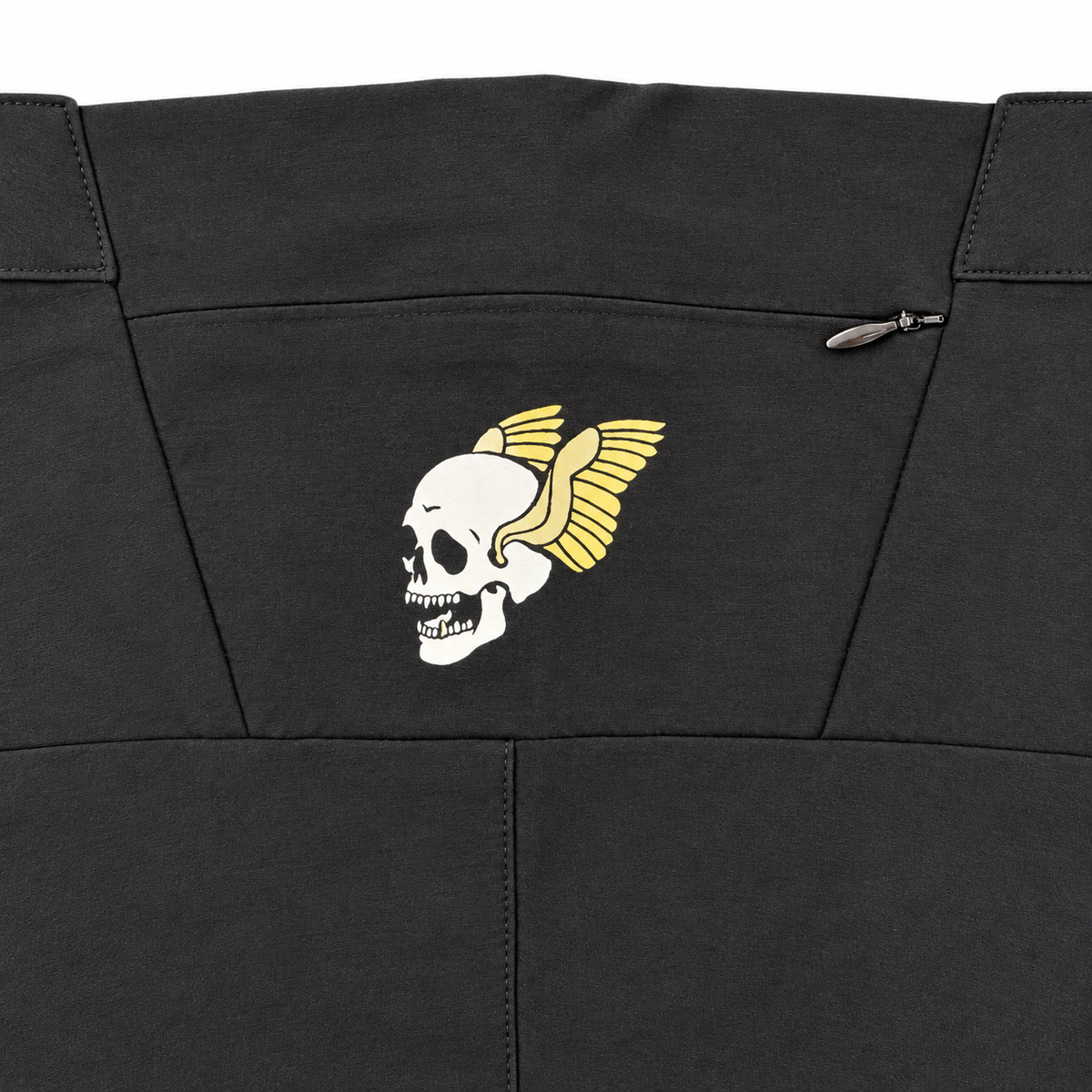 Close-up of a black garment featuring a white skull with yellow wings embroidered on the chest, with a small zipper pocket on the right side.