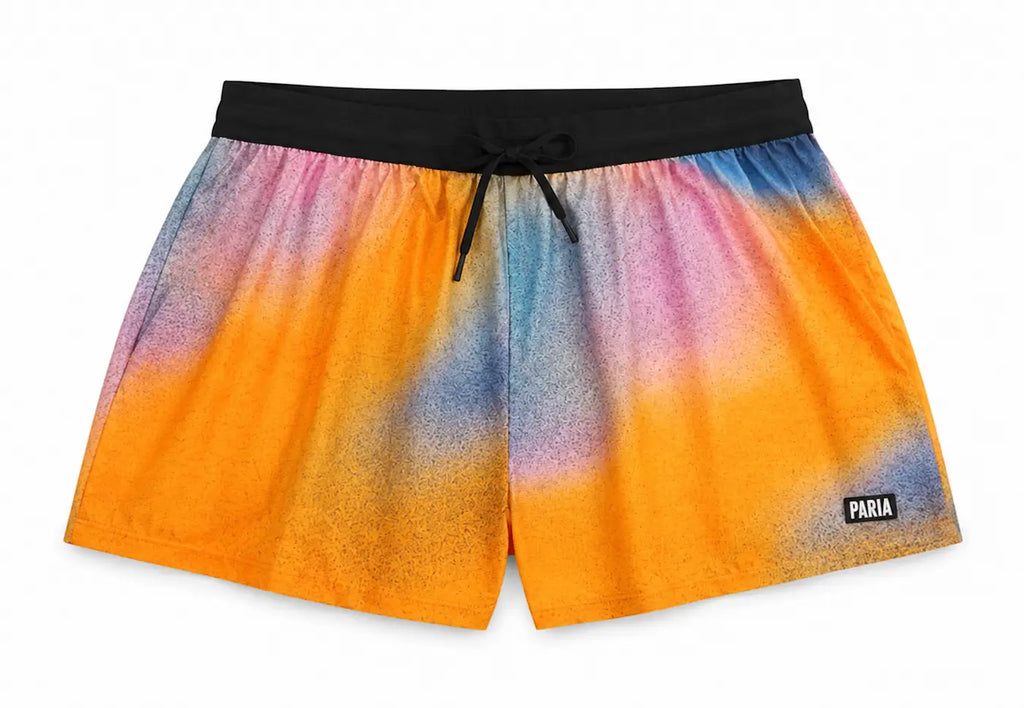 A pair of PARIA brand swim trunks featuring a vibrant orange, yellow, pink, and blue tie-dye gradient pattern, a black elastic waistband, and a black drawstring, shown against a plain white background.