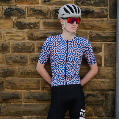 Person wearing a cycling outfit with a helmet and sunglasses against a brick wall.