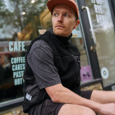 Man sitting outside a cafe wearing a black gilet and red cap.