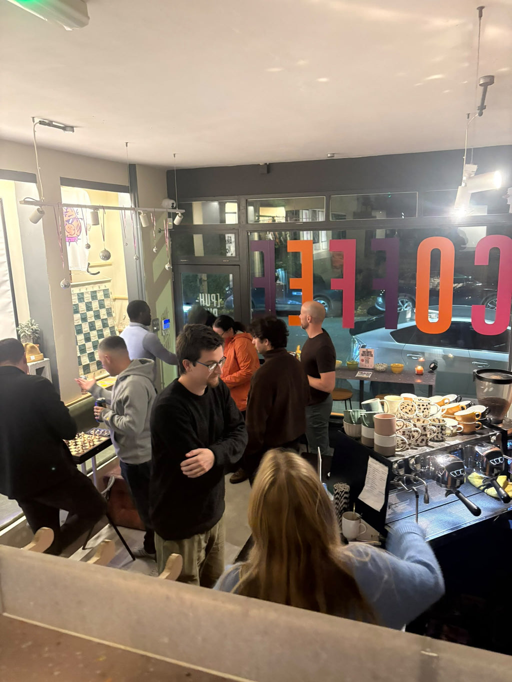 A busy cafe interior with several people standing and ordering near a counter, large colorful 'COFFEE' lettering on the window, and a glass display case holding pastries.