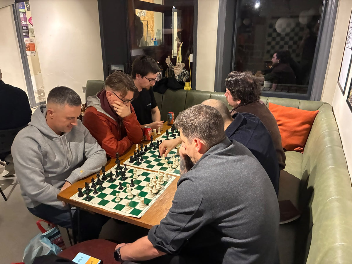 Several men gathered around a chessboard in a cozy indoor setting, with a green leather booth and orange pillows in the background.