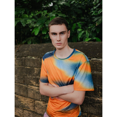 Person wearing a tie-dye shirt standing against a brick wall with greenery in the background
