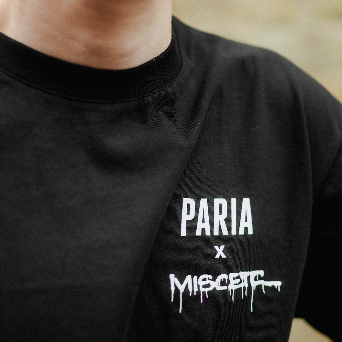Black t-shirt with 'PARIA x MISC ETC' text on a blurred natural background