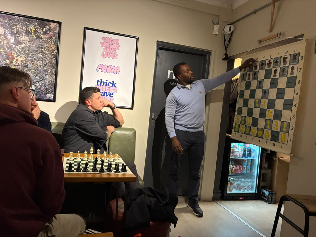 A man in a light blue sweater points to a large chessboard hanging on the wall in a room with other people seated nearby, including one at a chess table in the foreground.