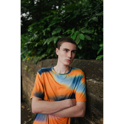 Person wearing a tie-dye shirt leaning against a stone wall with greenery in the background