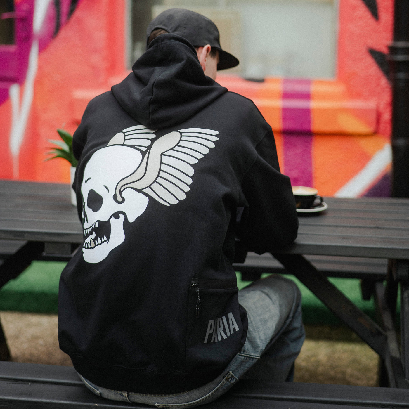 Person wearing a black hoodie with a skull design, sitting on a bench in front of a colorful mural.