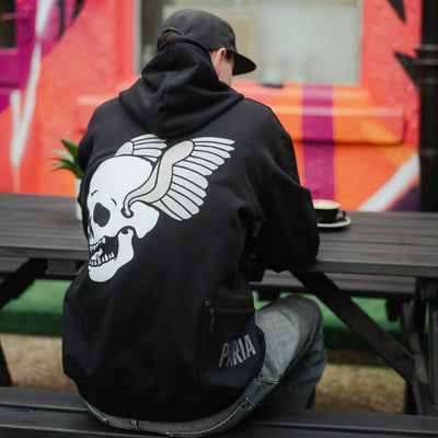 Person wearing a black hoodie with a skull design, sitting on a bench in front of a colorful mural.