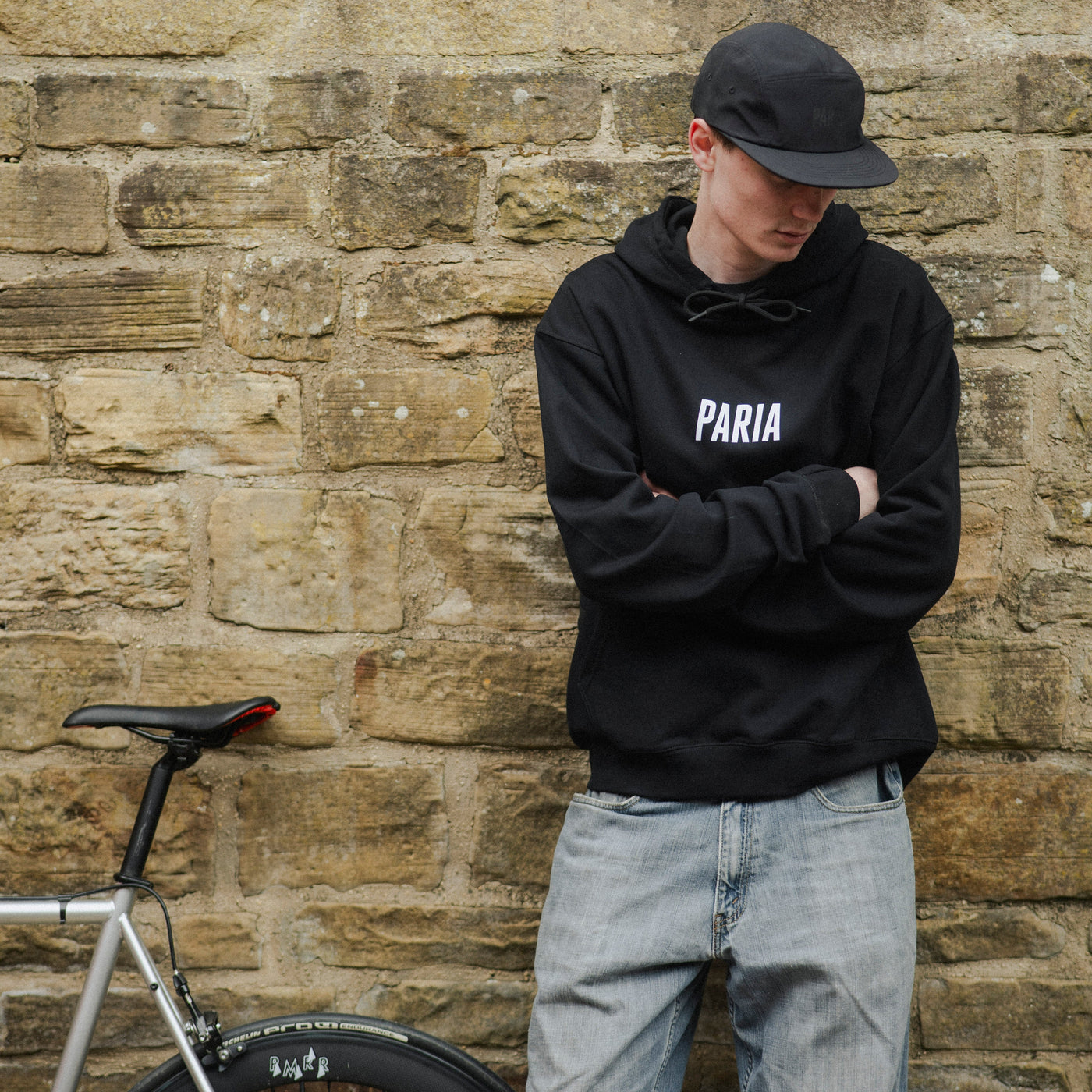 Person wearing a black hoodie with 'PARIA' branding against a stone wall with a bicycle.