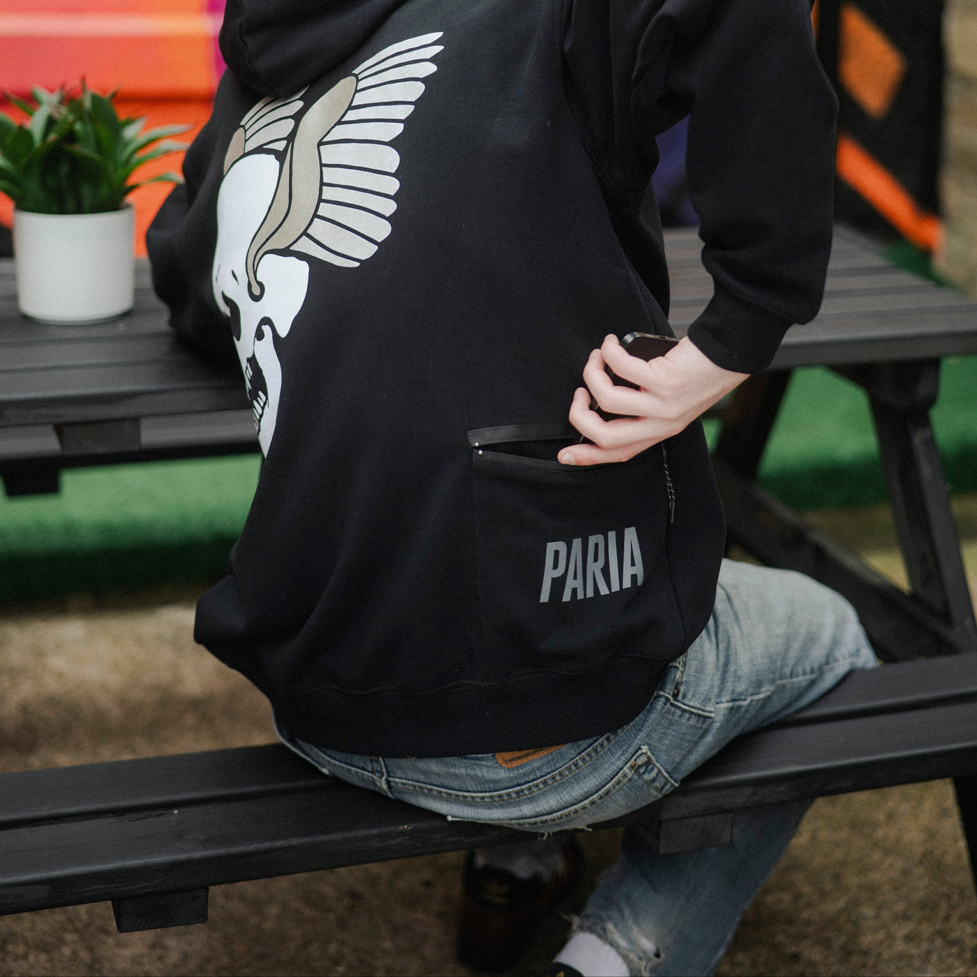 Person wearing a black hoodie with a winged logo and 'PARIA' branding, sitting on a bench.