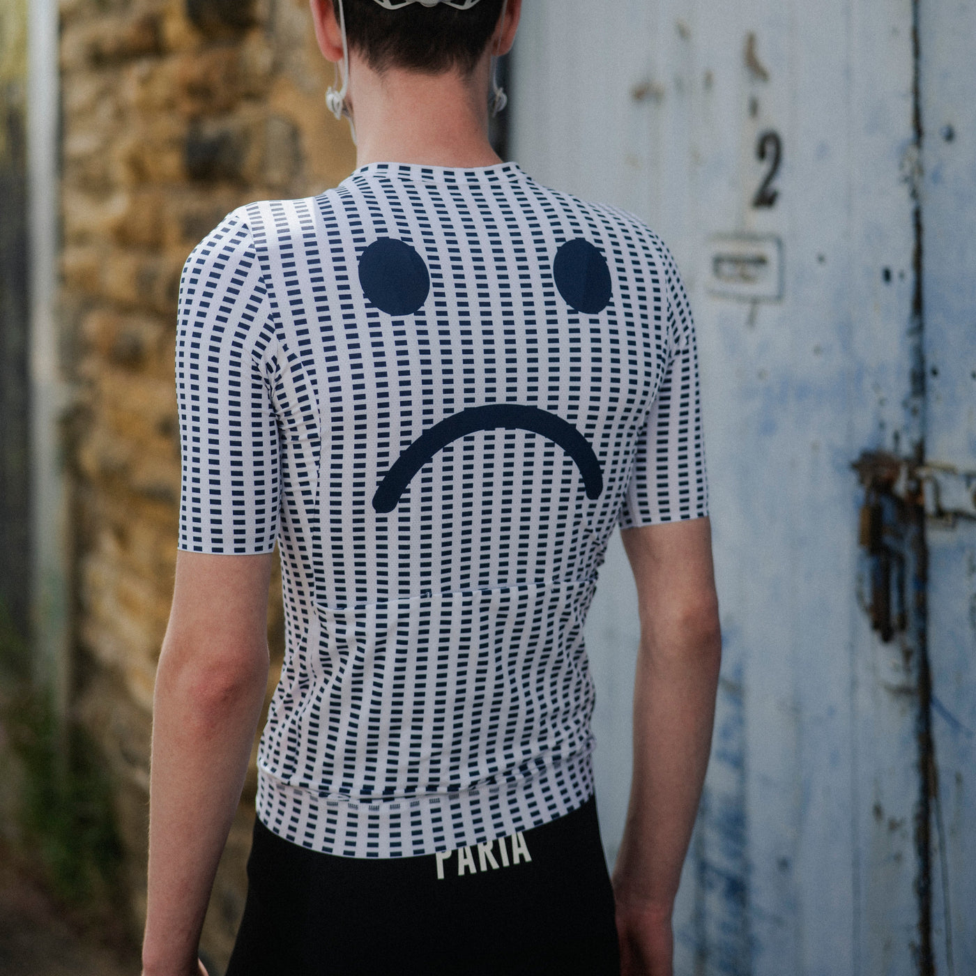 Person wearing a cycling helmet and a white shirt with a sad face design, standing in front of a textured wall.