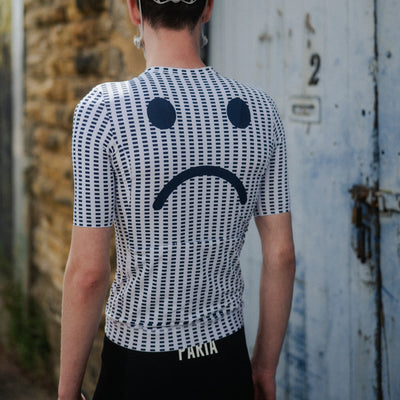 Person wearing a cycling helmet and a white shirt with a sad face design, standing in front of a textured wall.