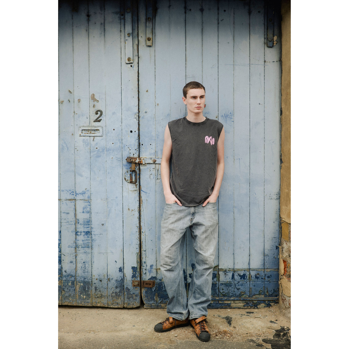 stone wash black sleeveless shirt with Paria pink logo and jeans standing in front of a textured wall.