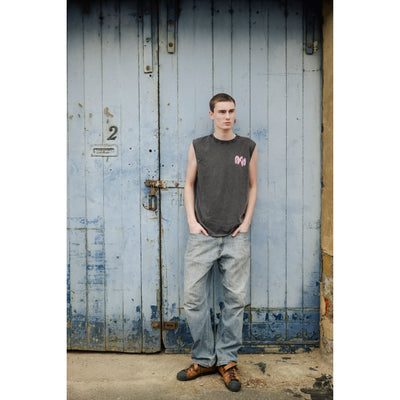 stone wash black sleeveless shirt with Paria pink logo and jeans standing in front of a textured wall.