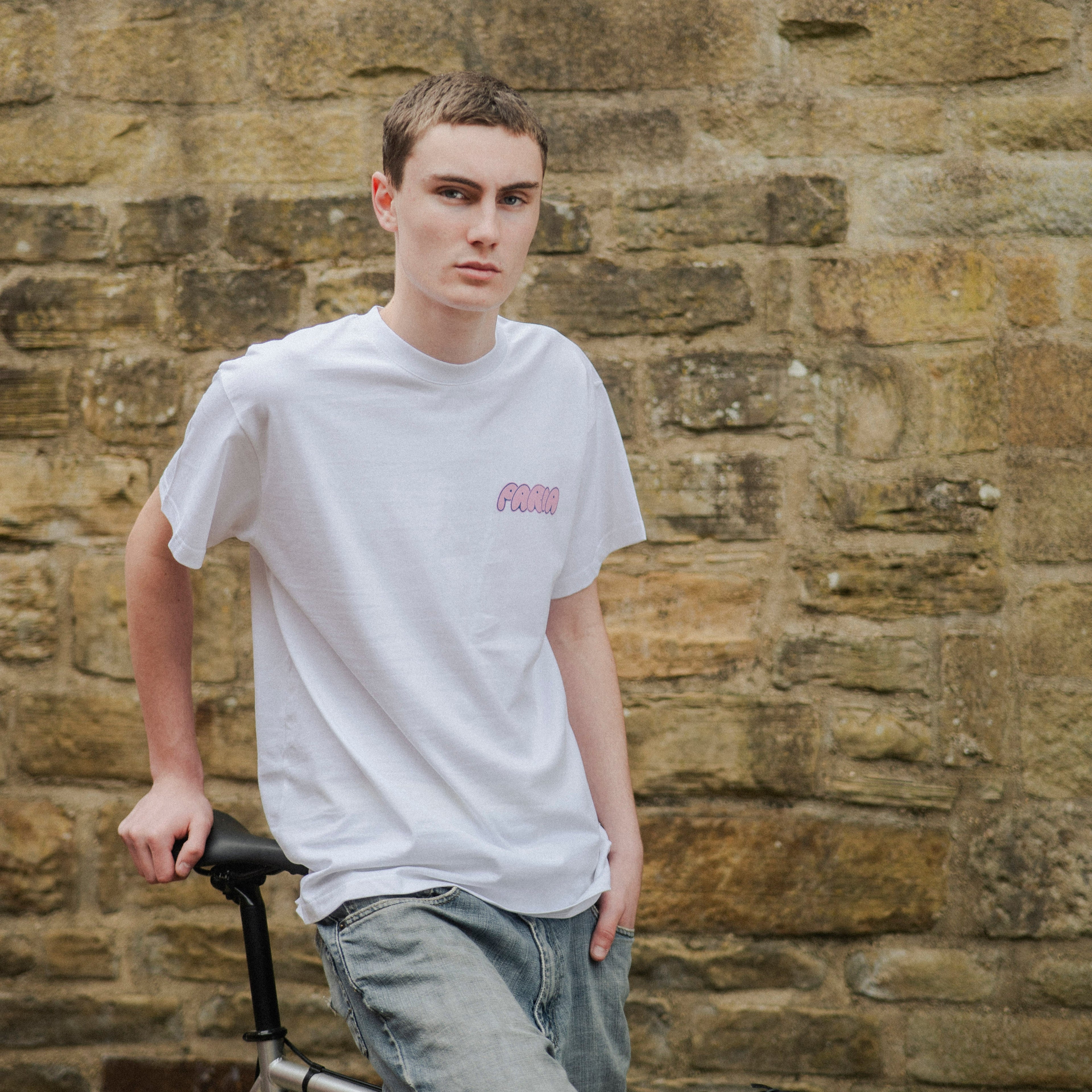 Person wearing a white t-shirt with a logo, standing next to a bicycle against a stone wall.