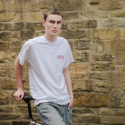 Person wearing a white t-shirt with a logo, standing next to a bicycle against a stone wall.