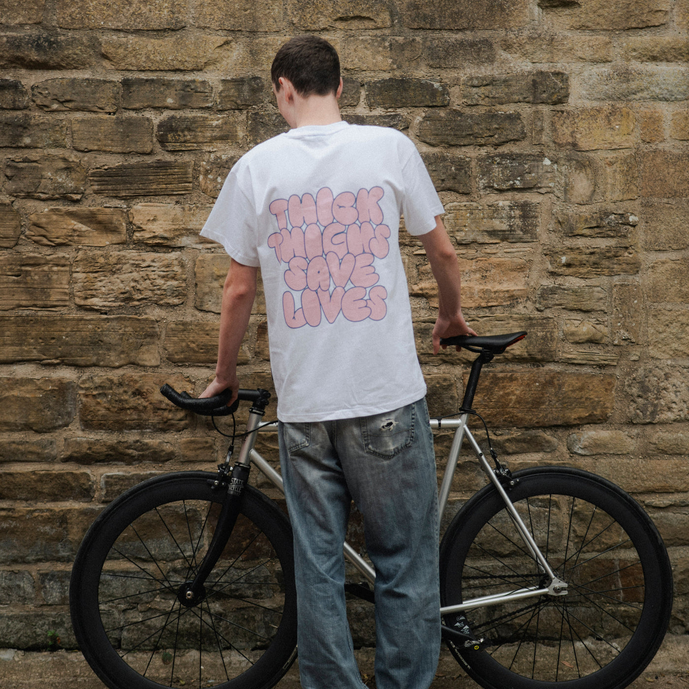 Person holding a bicycle against a stone wall wearing a white t-shirt with text.
