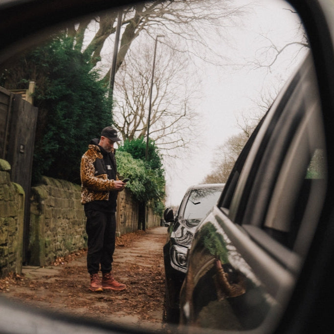 Person standing in leopard fleece on a path reflected in a car's side mirror