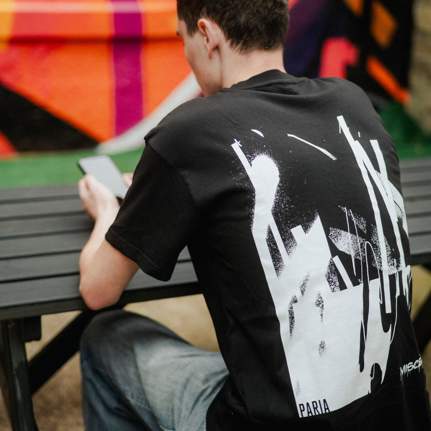 Person wearing a black t-shirt with white graphic design sitting at a table outdoors.