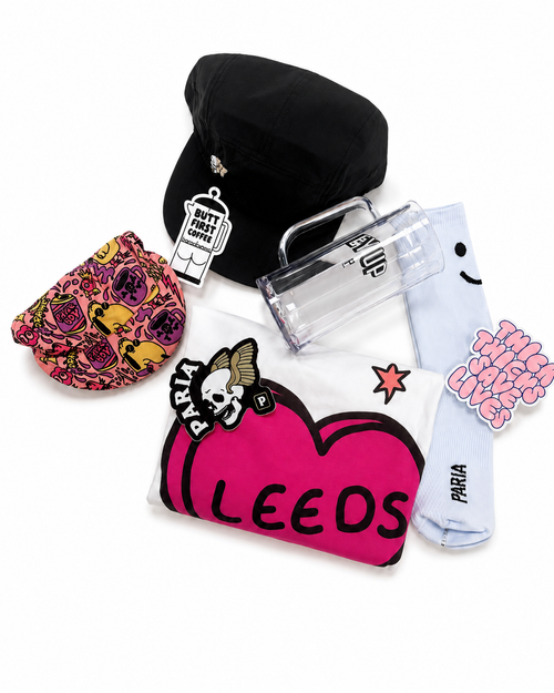 A curated flat-lay of fashion accessories from the brand Faria, including a black cap with a Doctor Who badge, a pink and yellow floral patterned pouch, a clear plastic tumbler, a white graphic T-shirt with a large pink heart and 'LEEDS' text, and a light blue pair of socks with 'THIS IS MY LIFE' text.