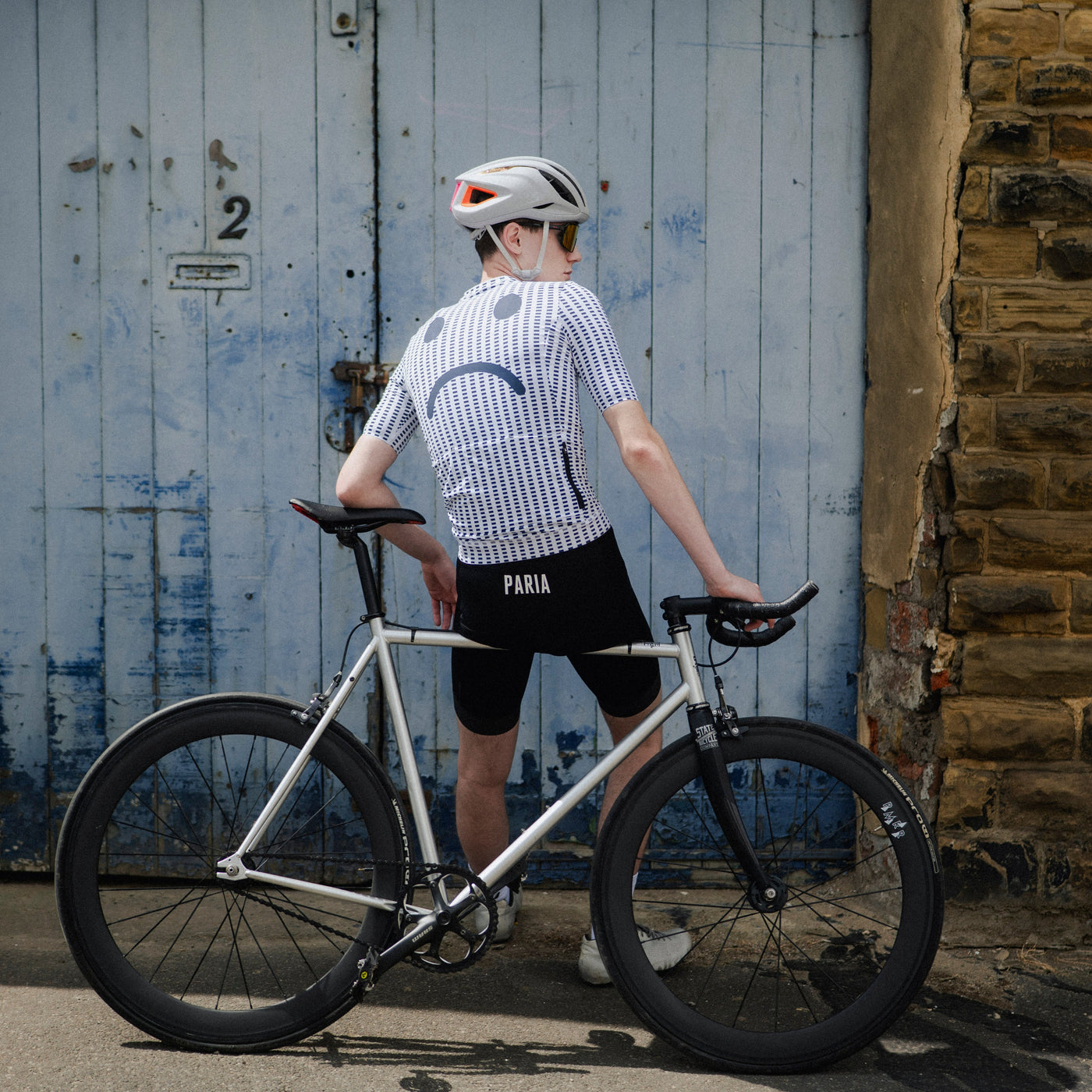 Person wearing a cycling helmet and a white shirt with a sad face design, standing in front of a textured wall.