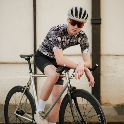 Person sitting on a bicycle wearing a helmet and sunglasses against a plain wall.