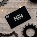 A black gift card with the name 'PARIA' printed in white, surrounded by bicycle parts like gears and a chain, suggesting a cycling theme.